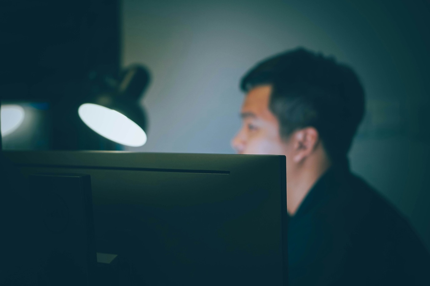 A person sits in profile at a softly lit monitor in a darkened workspace