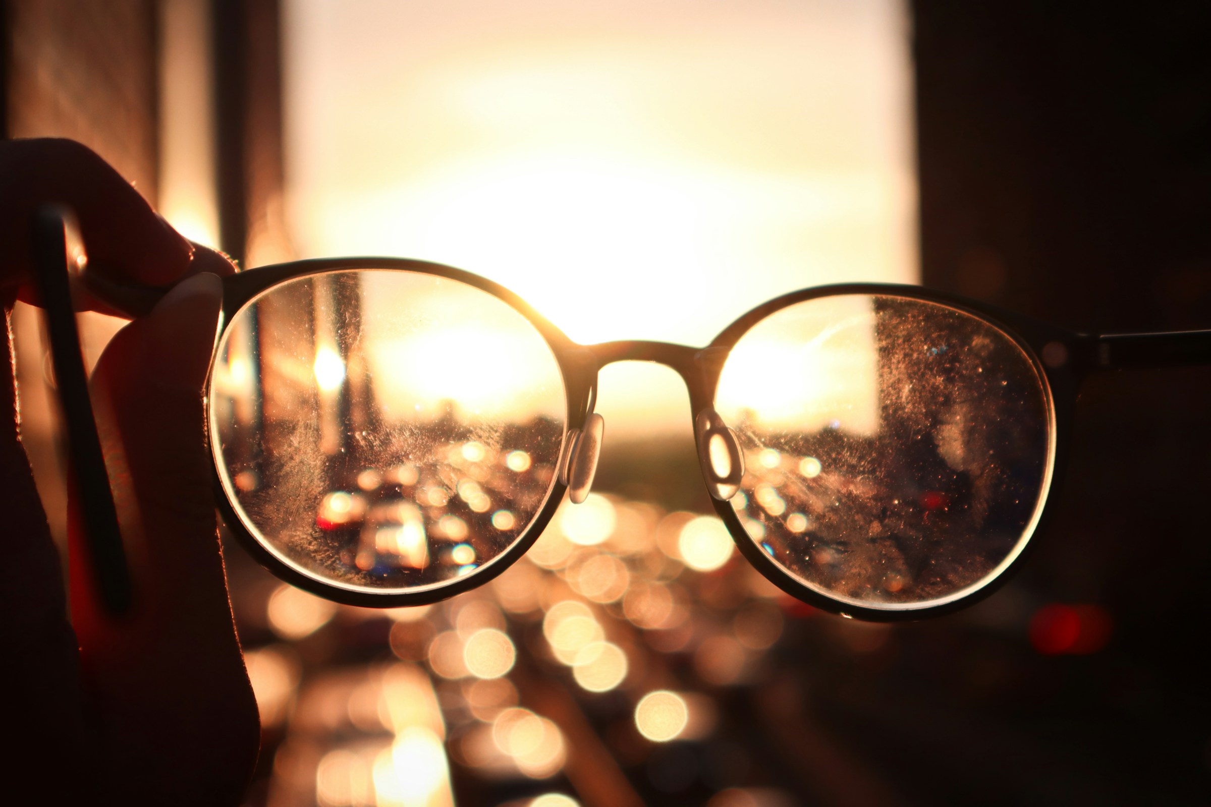 A hand holds eyeglasses up to a blurred city sunset, bringing the scene into sharp focus through the lenses
