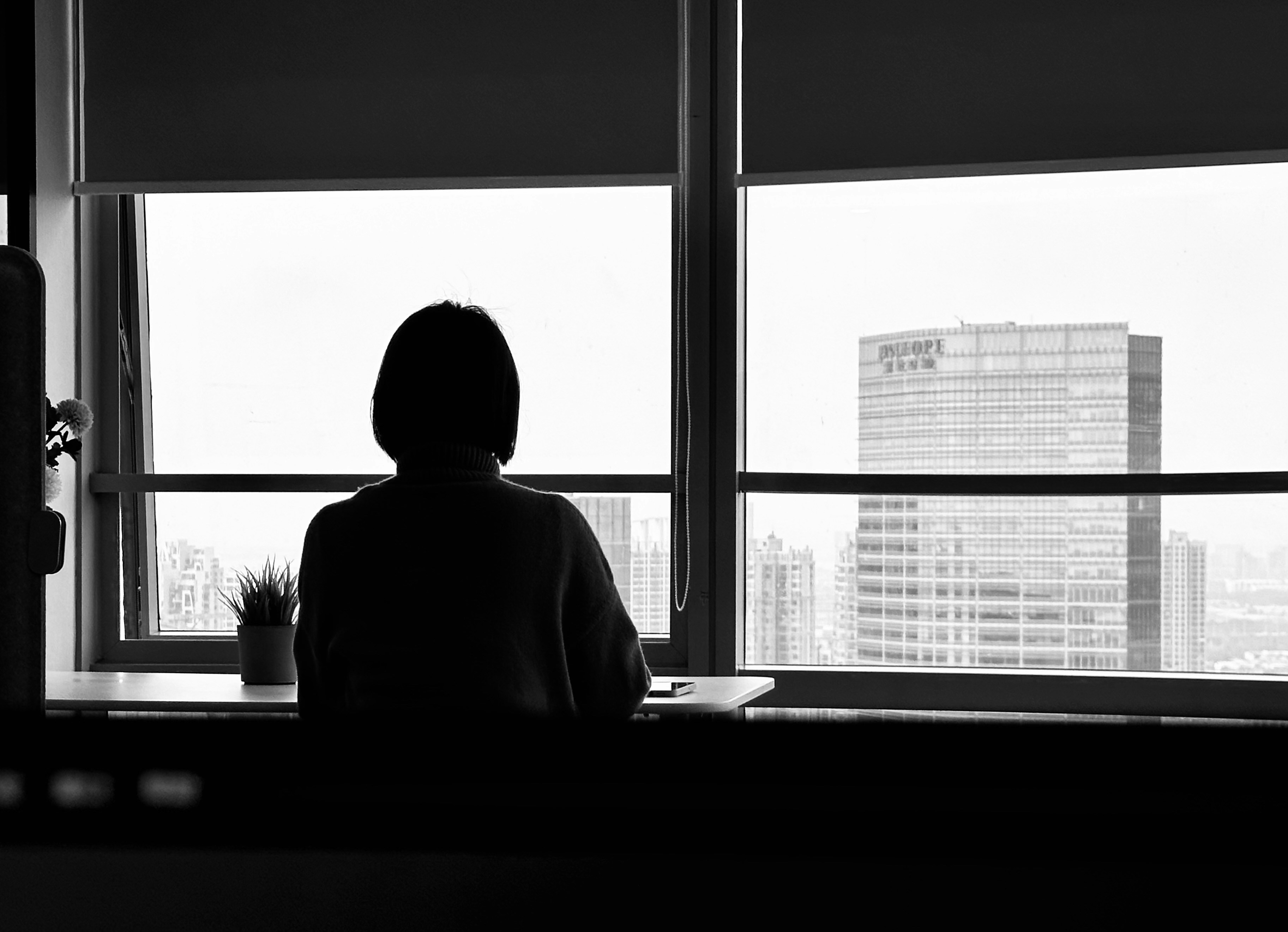 A person sits alone in silhouette at a high-rise office window, looking out over a city skyline
